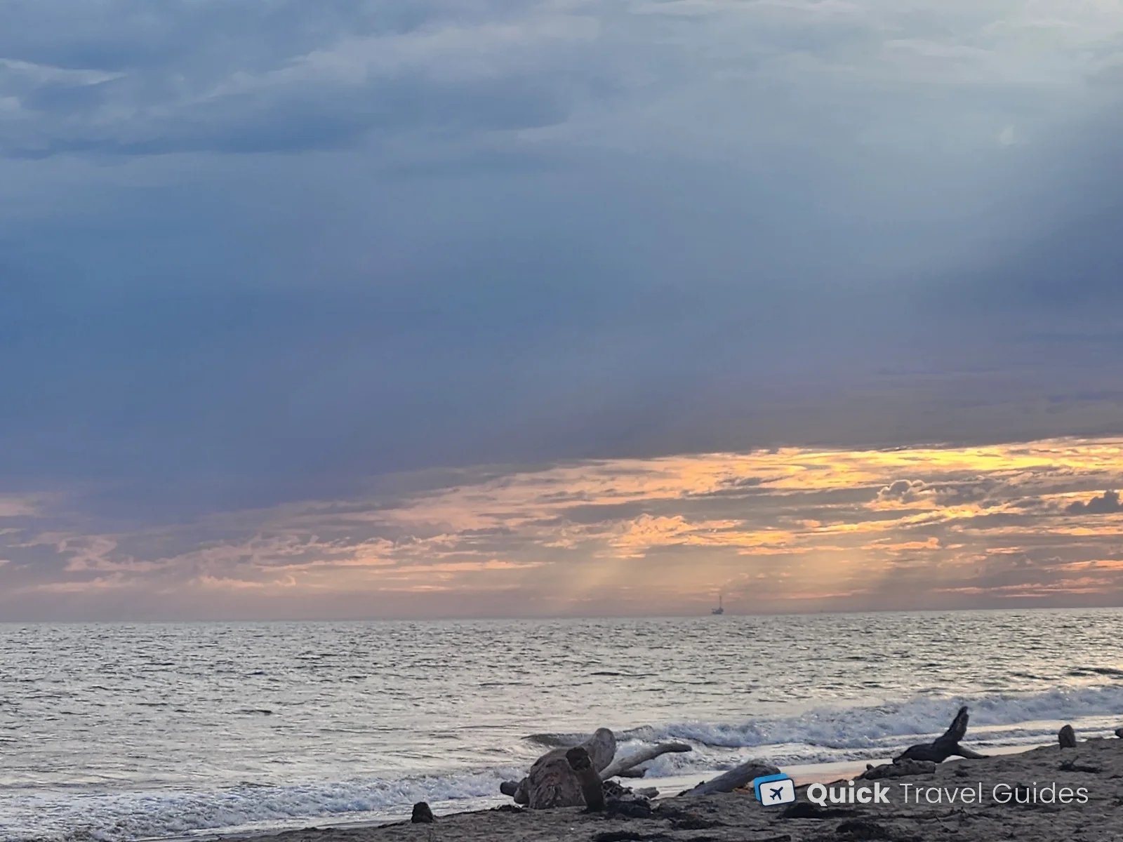 An evening sky over the Santa Barbara Channel, with driftwood on the beach and an oil platform on the horizon