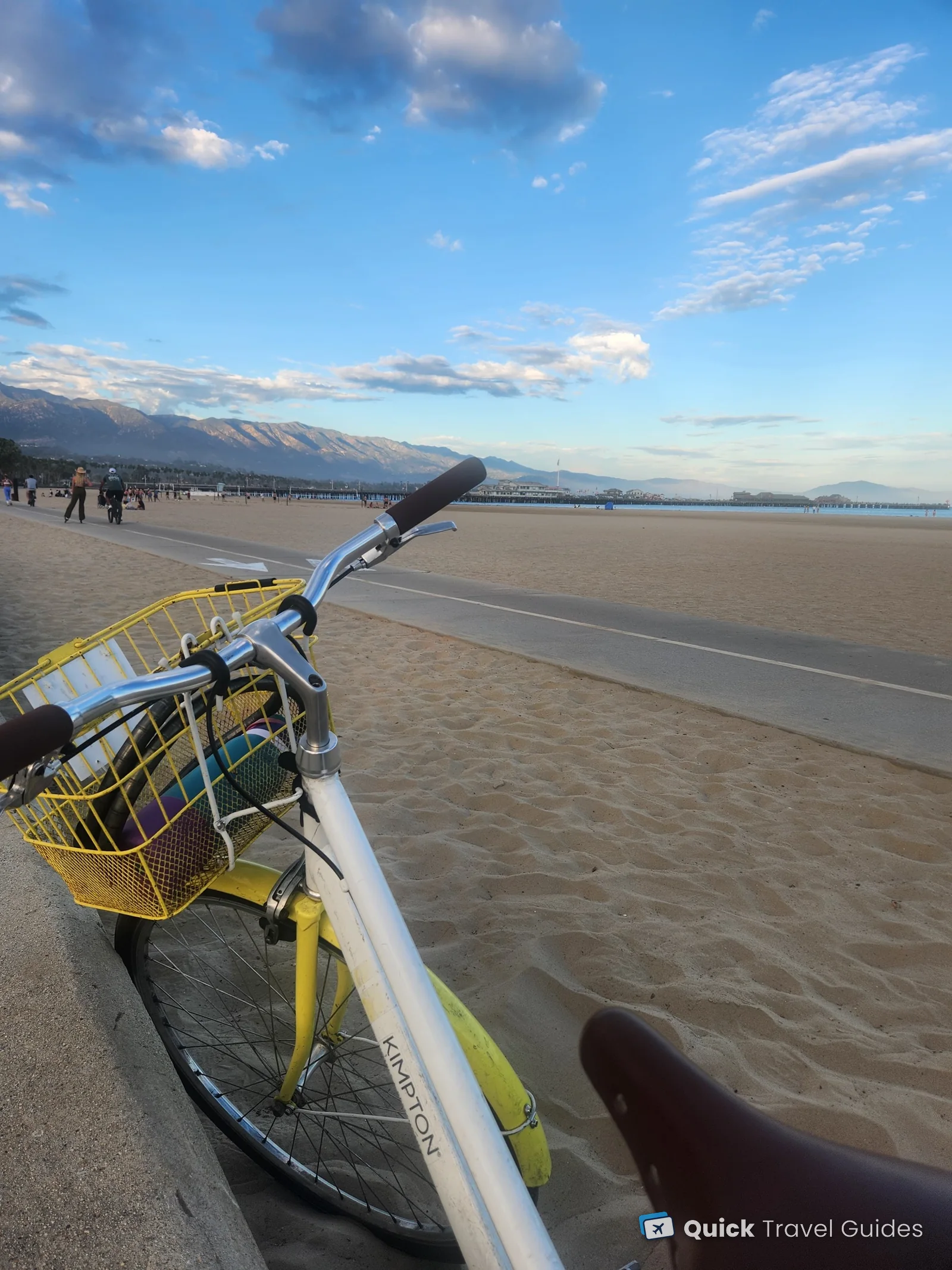 A Kimpton Canary bike with a yellow basket on the beachfront bike path in Santa Barbara, with Stearns Wharf and the Santa Ynez Mountains behind