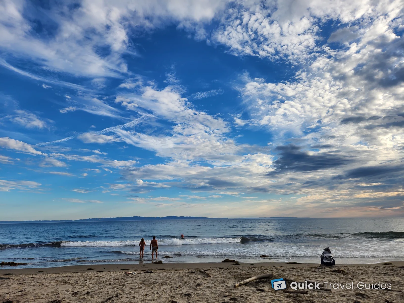 Swimmers at the water's edge on a Santa Barbara beach, with the Channel Islands on the horizon under a big sky